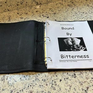 Op de afbeelding: Open zwarte map met de titel "Bound By Bitterness" op een witte pagina. De pagina bevat een zwart-wit afbeelding van in elkaar geslagen handen en kettingen. De map is open op een gespikkelde aanrecht.