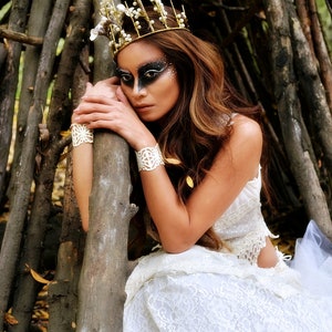 May include: A woman wearing a white lace dress and a gold crown with white flowers. She has dark hair and dark makeup with white accents. She is posing with her hands on a tree branch.