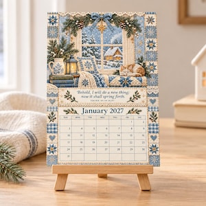 Puede incluir: Un calendario de enero de 2027 sobre un caballete de madera con una escena invernal, un gato, una ventana nevada y libros. El texto dice: Behold, I will do a new thing; now it shall spring forth. Isaiah 43:19 (KJV). Borde con patrones azules.