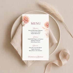 Op de afbeelding: Een trouwmenu met bloemversieringen, met voorgerechten, hoofdgerechten en desserts. Het menu bevat gerechten zoals geroosterde rode paprika hummus en veganistische cheesecake. De namen "Carmen & Andrew" staan onderaan afgedrukt.