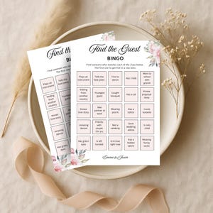 Op de afbeelding: Twee witte Bingokaarten met bloemaccenten en de tekst "Find the Guest Bingo". De kaarten hebben een raster met prompts zoals "Heeft een tatoeage" en "Houdt van karaoke". De kaarten liggen op een witte schaal.