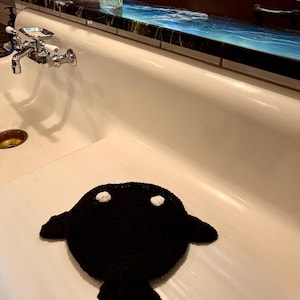 May include: A circular black crocheted textile piece featuring two small white circular accents near the top edge, two side protrusions, and a bottom tail-like extension, creating a stylized animal-like shape with a textured yarn surface.