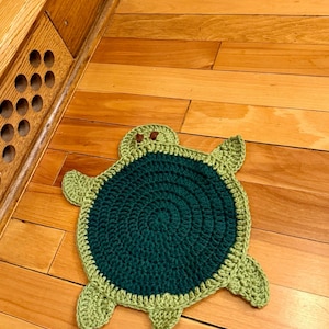 May include: A handmade crochet turtle-shaped mat featuring a dark forest green circular center and a light lime green border, head, and four flippers. The turtle has two small brown stitched eyes on its head, creating a flat, decorative piece.
