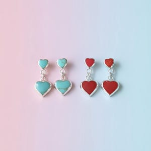 May include: Silver heart-shaped dangle earrings. Two pairs feature turquoise and red heart-shaped stones. The earrings have a small heart at the top and a larger heart at the bottom, connected by a silver chain. The background is a gradient of pink and blue.
