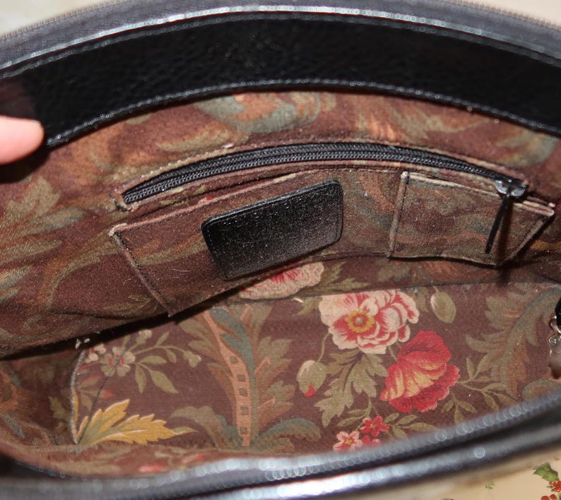 May include: Open view of a black leather handbag interior. The inside features a floral fabric lining with red and pink flowers, green leaves, and a brown background. There are several pockets and a zippered compartment.