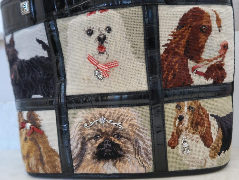 May include: A tapestry tote bag featuring various dog breeds. The bag has a black leather trim and handles. The tapestry panels depict different dogs, including a white dog with a red bow, and a brown and white dog.
