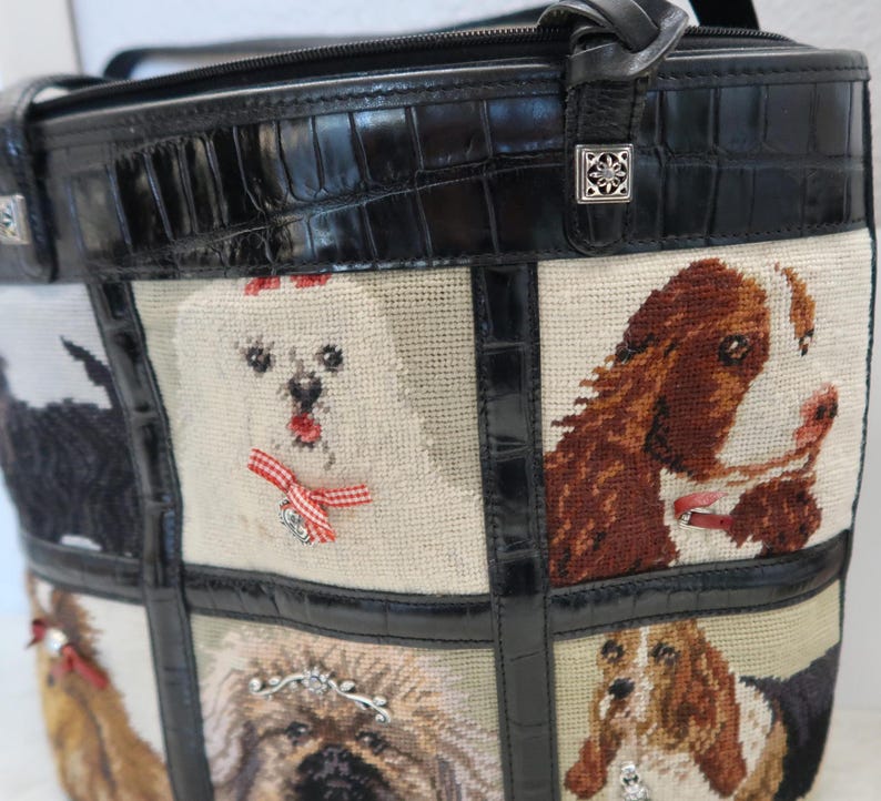 May include: A black tote bag with a stitched dog design. The bag features a black crocodile-embossed leather top and trim. The dog portraits are on a beige background, with red bows and silver embellishments.