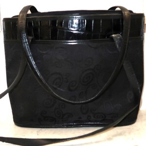 May include: Black tote bag with a textured, embossed pattern. The bag features a glossy, faux-crocodile top section and long, slender handles. A detachable shoulder strap is also included.