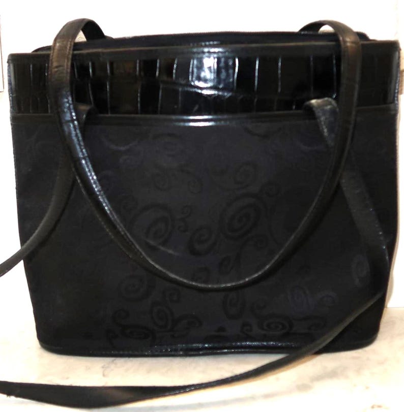May include: Black tote bag with a textured, embossed pattern. The bag features a glossy, faux-crocodile top section and long, slender handles. A detachable shoulder strap is also included.