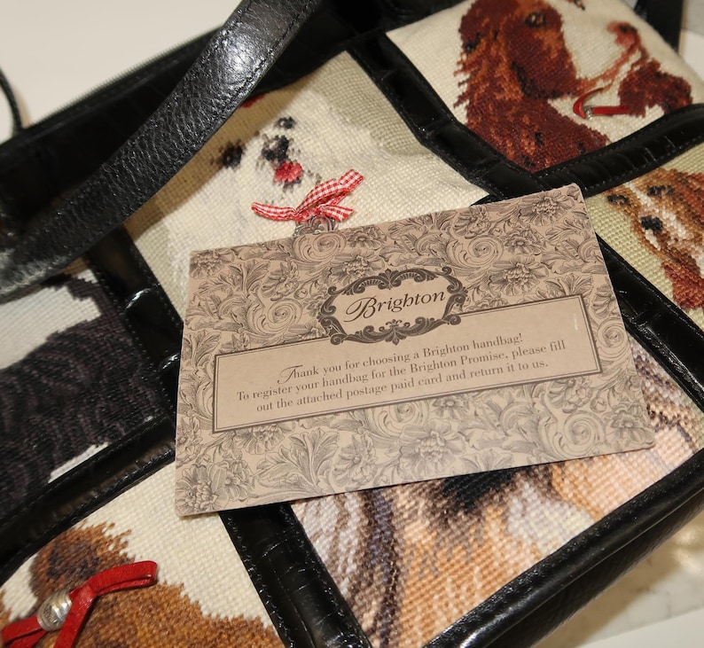 May include: A black leather handbag with a patchwork design featuring embroidered dog portraits. The bag has a top handle and a decorative card that reads "Brighton". The card includes text about registering the handbag.