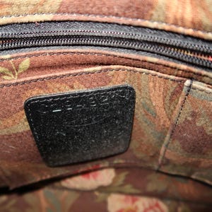 May include: Close-up of a handbag interior with a black zipper and a black leather tag. The interior fabric features a floral pattern in shades of brown, green, and red. The tag is rectangular and stitched into the fabric.