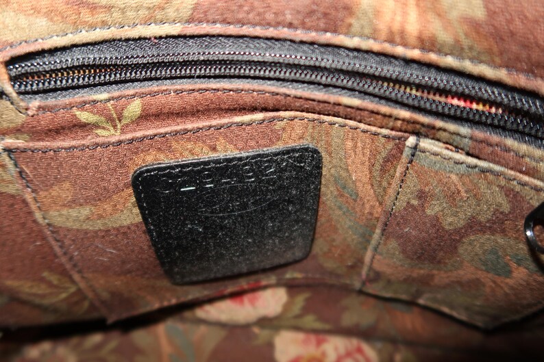 May include: Close-up of a handbag interior with a black zipper and a black leather tag. The interior fabric features a floral pattern in shades of brown, green, and red. The tag is rectangular and stitched into the fabric.