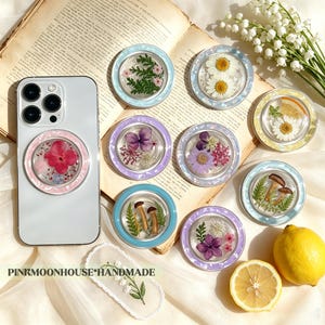 May include: A collection of circular phone grips featuring pressed flowers, mushrooms, and greenery encased in resin with pastel-coloured borders. One grip is attached to a white smartphone. The text PINKMOONHOUSE*HANDMADE is visible at the bottom.