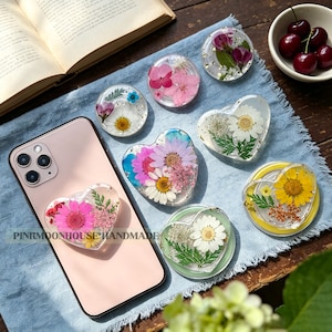May include: A collection of circular and heart-shaped phone grips made of clear resin, each embedded with various pressed flowers and green foliage. One heart-shaped grip is attached to a light pink smartphone with the text PINKMOONHOUSEHANDMADE.