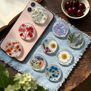 May include: A collection of circular phone grips featuring clear resin embedded with various pressed flowers, including white baby's breath, red flowers, orange blossoms, blue forget-me-nots, daisies, and green ferns, arranged on a light blue cloth.