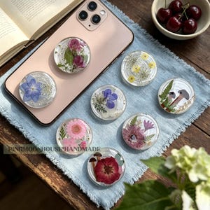 May include: A collection of eight circular phone grips featuring clear resin embedded with pressed flowers, leaves, and mushrooms. The designs include pink, blue, purple, and red floral arrangements. One grip is attached to a rose gold smartphone.