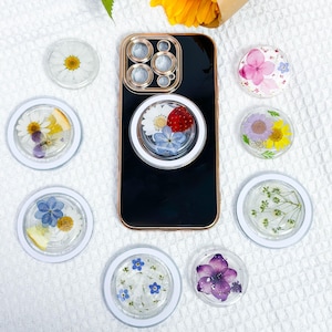 Könnte beinhalten: Ein schwarzes Handy-Case mit einem goldenen Ringhalter und einem durchsichtigen Handy-Griff mit gepressten Blumen. Der Handy-Griff hat eine weiße Margerite, blaue Vergissmeinnicht und eine rote Erdbeere. Es gibt acht kleinere durchsichtige Handy-Griffe mit gepressten Blumen in verschiedenen Farben und Designs.