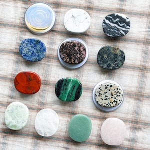 May include: Assortment of twelve gemstone phone grips in various colors and patterns. Includes grips with blue, white, black, red, green, and pink stones. Each grip is round or oval and set in a silver-colored base.
