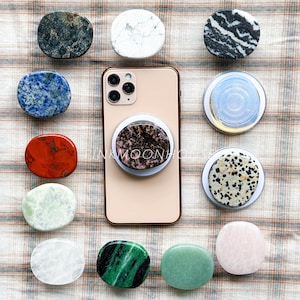 May include: A selection of gemstone phone grips in diverse colours and patterns, including marble and jasper. A phone with a pink and brown stone grip is in the centre. The grips are oval and circular.