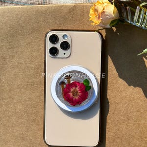 Può includere: Uno smartphone beige con un supporto trasparente per telefono con una rosa rossa essiccata, due piccoli funghi e foglie verdi. Il supporto del telefono ha il testo "PINKMOONHOUSE" in bianco.
