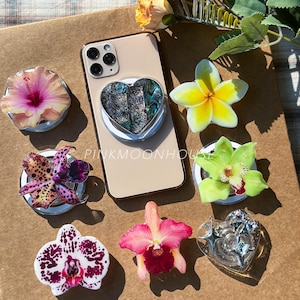 Op de afbeelding: Een verzameling bloemen telefoonhouders en een telefoon met een hartvormige houder. De houders hebben verschillende bloemmotieven, waaronder hibiscus, orchideeën en frangipani. De telefoon heeft een goudkleurige achterkant.