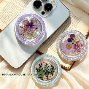 Peut inclure: Trois supports de téléphone circulaires avec des fleurs séchées sous résine transparente et des bordures nacrées. Un support est fixé sur un smartphone blanc, tandis que deux autres présentent des compositions florales violettes, roses et vertes. Texte : PINKMOONHOUSE*HANDMADE.