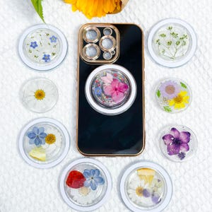 May include: Assortment of phone grips with pressed flowers and leaves. Each grip features a different floral design, including blue forget-me-nots, white daisies, pink hydrangeas, purple flowers, and a mix of yellow and purple flowers.