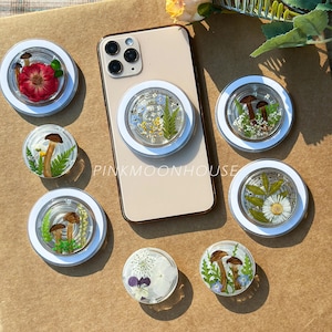 May include: A collection of round phone grips with various designs. Some grips feature dried flowers and greenery, while others showcase mushroom illustrations. One grip is attached to a gold phone. The text "PINKMOONHOUSE" is visible.