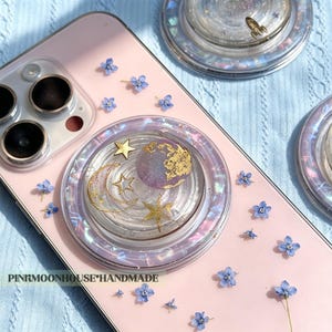 May include: A light pink smartphone case decorated with small blue pressed flowers and a circular phone grip featuring a purple and gold planet, a crescent moon, and stars. The text PINKMOONHOUSE*HANDMADE is printed on the lower left side.