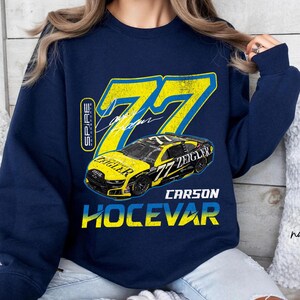 May include: Navy blue sweatshirt with a yellow and blue race car graphic, displaying the number 77. The design includes the name "Carson Hocevar" and the text "Zeigler Cars". The sweatshirt also features the word "Spire" on the left side.