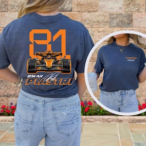 May include: A navy blue t-shirt featuring a Formula 1 race car design with the number 81 in orange. The shirt also displays the name "Oscar Piastri" and the McLaren logo. The design is on the back of the shirt.