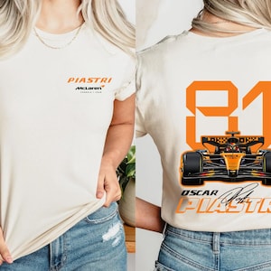 Puede incluir: Camiseta color crema con el nombre "PIASTRI" en naranja y el logo de McLaren. La parte trasera muestra un gran "81" naranja con un gráfico de coche de Fórmula 1 y el texto "OSCAR PIASTRI".