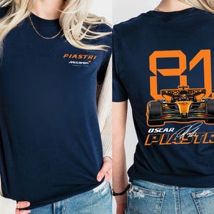 May include: Navy blue t-shirt featuring the name "PIASTRI" and "McLaren" in orange on the front. The back displays the number "81" and a Formula 1 car graphic, along with "OSCAR PIASTRI" in orange.