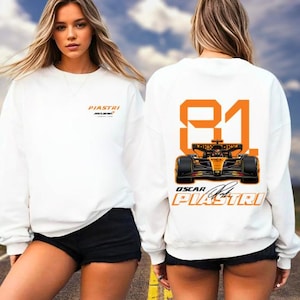 May include: White sweatshirt featuring the name "PIASTRI" and the McLaren logo in orange. The back displays the number "81" and a race car graphic in orange and black, along with "OSCAR PIASTRI" and a signature.