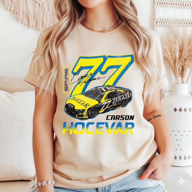 May include: Beige t-shirt with a yellow and blue graphic. The design includes the number 77, a race car, and the name "Carson Hocevar". The shirt also has the text "Spire" and "Zeigler".