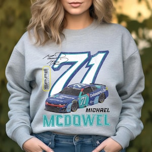 May include: A light gray sweatshirt featuring a racing car design. The design includes the number 71, a blue race car, the name "MICHAEL MCDOWEL", and the "Delaware Life" logo. The sweatshirt also has a signature and the "SPIRE MOTORSPORTS" logo.