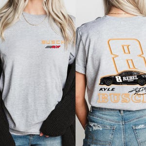 Puede incluir: Camiseta gris con un gr&aacute;fico de coche de carreras negro y el texto "Kyle Busch" y "8 Rebel" en la espalda. La parte delantera de la camiseta tiene el texto "Busch" en rojo y naranja.