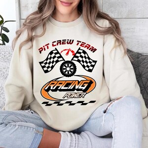Puede incluir: Sudadera color crema con un diseño temático de carreras. El diseño incluye el texto "PIT CREW TEAM" en rojo, banderas a cuadros, un velocímetro, un neumático y las palabras "RACING POWER" en un óvalo naranja y negro.