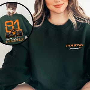 May include: Dark green sweatshirt featuring the name "PIASTRI" in orange above the McLaren logo. The back of the sweatshirt has the number "81" in orange, a race car graphic, and the name "OSCAR PIASTRI".