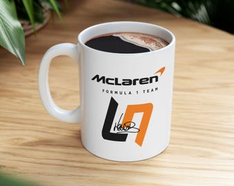 May include: White ceramic mug with the McLaren Formula 1 Team logo and a black and orange design. The mug is filled with dark coffee. The mug has a white handle and is sitting on a wooden surface.