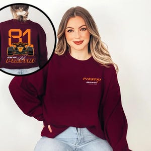 May include: Burgundy sweatshirt featuring the name "PIASTRI" in orange, with the McLaren logo below. The back of the sweatshirt displays the number "81" and a race car graphic in orange and black, with "OSCAR PIASTRI" below.