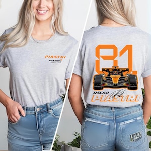May include: A light gray t-shirt featuring the name "PIASTRI" and "McLaren" in orange. The back of the shirt displays the number "81" in orange, a race car graphic, and the name "OSCAR PIASTRI".