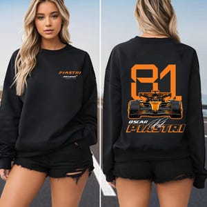 May include: Black sweatshirt featuring the name "PIASTRI" and "McLaren" in orange. The back displays the number "81" and a race car graphic in orange, along with "OSCAR PIASTRI" and a signature. The sweatshirt is worn by a person.