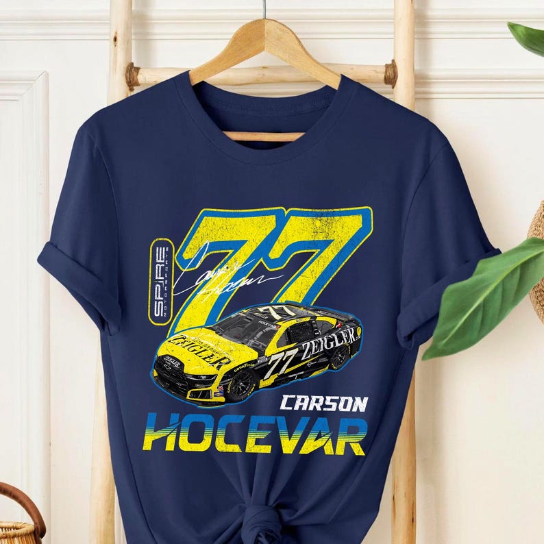May include: Navy blue t-shirt with a yellow and black race car design and the number 77. The shirt features the name "Carson Hocevar" and the word "Zeigler". The t-shirt is displayed on a wooden hanger.