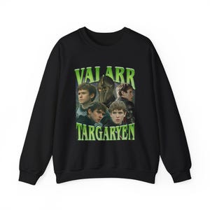 以下が含まれることがあります： 黒いスウェットシャツ。緑色のグラフィックで「VALARR TARGARYEN」と書かれており、ファンタジーシリーズのキャラクターの画像が添えられています。スウェットシャツはクルーネックです。