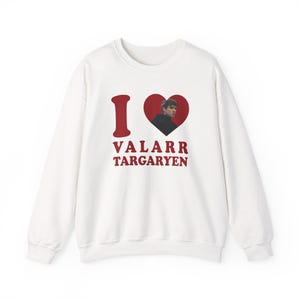 以下が含まれることがあります： 「I ❤️ VALARR TARGARYEN」の赤い文字が入った白いスウェットシャツ。ハートのグラフィックには人物の画像が含まれています。スウェットシャツはクルーネックです。