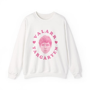 May include: White crew neck sweatshirt with a pink graphic. The graphic features a portrait of a person with pink hair, surrounded by the words "VALARR TARGARYEN" in a circle, with pink hearts. The sweatshirt is a casual style.