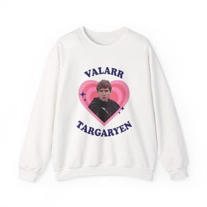 May include: White sweatshirt featuring a pink heart design with the text "VALARR TARGARYEN" in blue. The heart contains an image of a person. The sweatshirt is a casual garment.
