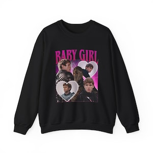 以下が含まれることがあります： 黒色のスウェットシャツ。ピンク色の「BABY GIRL」の文字と、ハート型のフレームに入った人物の画像がプリントされています。カジュアルで快適な衣服です。