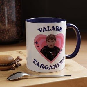 以下が含まれることがあります： 内側とハンドルが青い白いセラミックマグ。マグには「VALARR TARGARYEN」の文字と人物の画像が入ったピンクのハートのデザインが施されています。木製の表面にはスプーンとペストリーがあります。